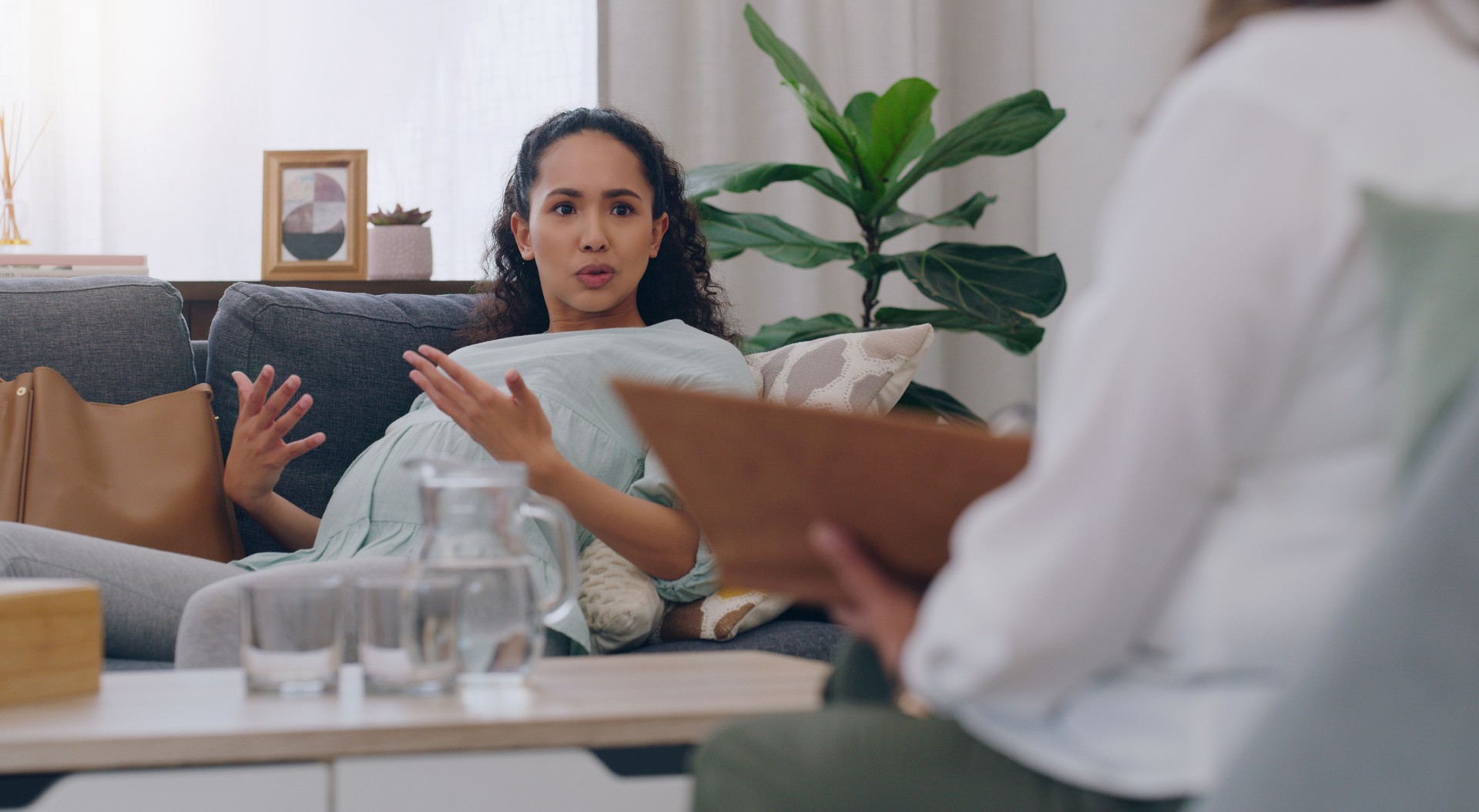 Stress, anxiety or pregnant woman talking to psychologist on mental health, support or perinatal depression healthcare clinic sofa. Pregnancy, fear or scared mother on couch in psychology counseling