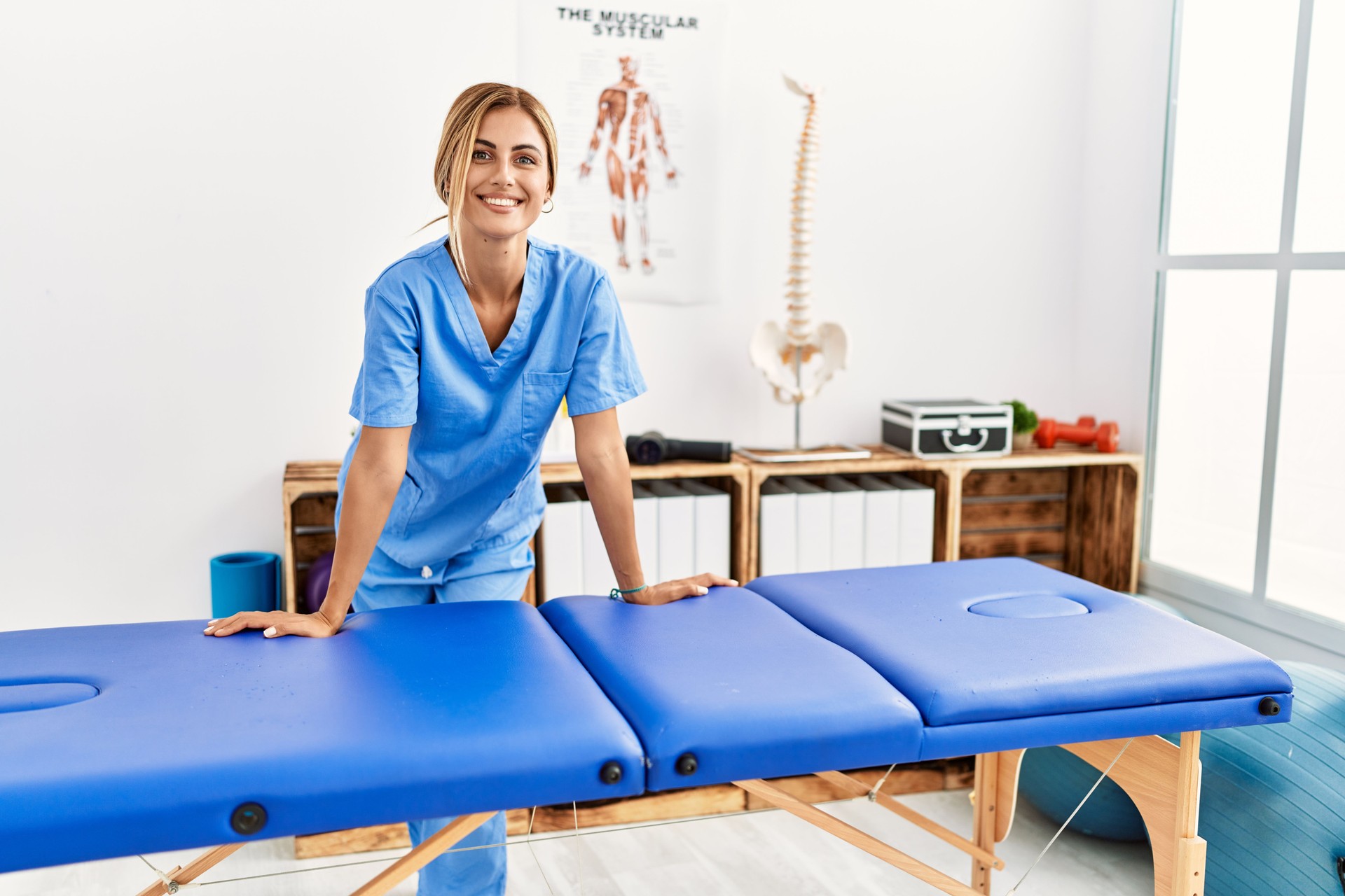 Young caucasian physio therapist smiling happy leaning on table at the clinic