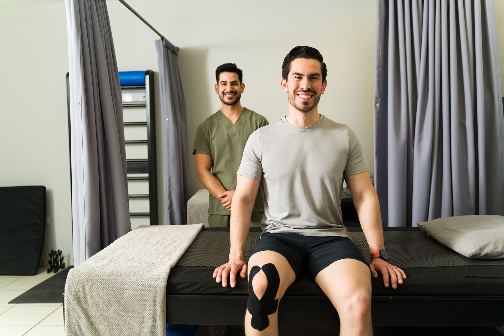 Man recovering from knee injury, sitting on massage table with kinesiology tape, smiling during physical therapy treatment