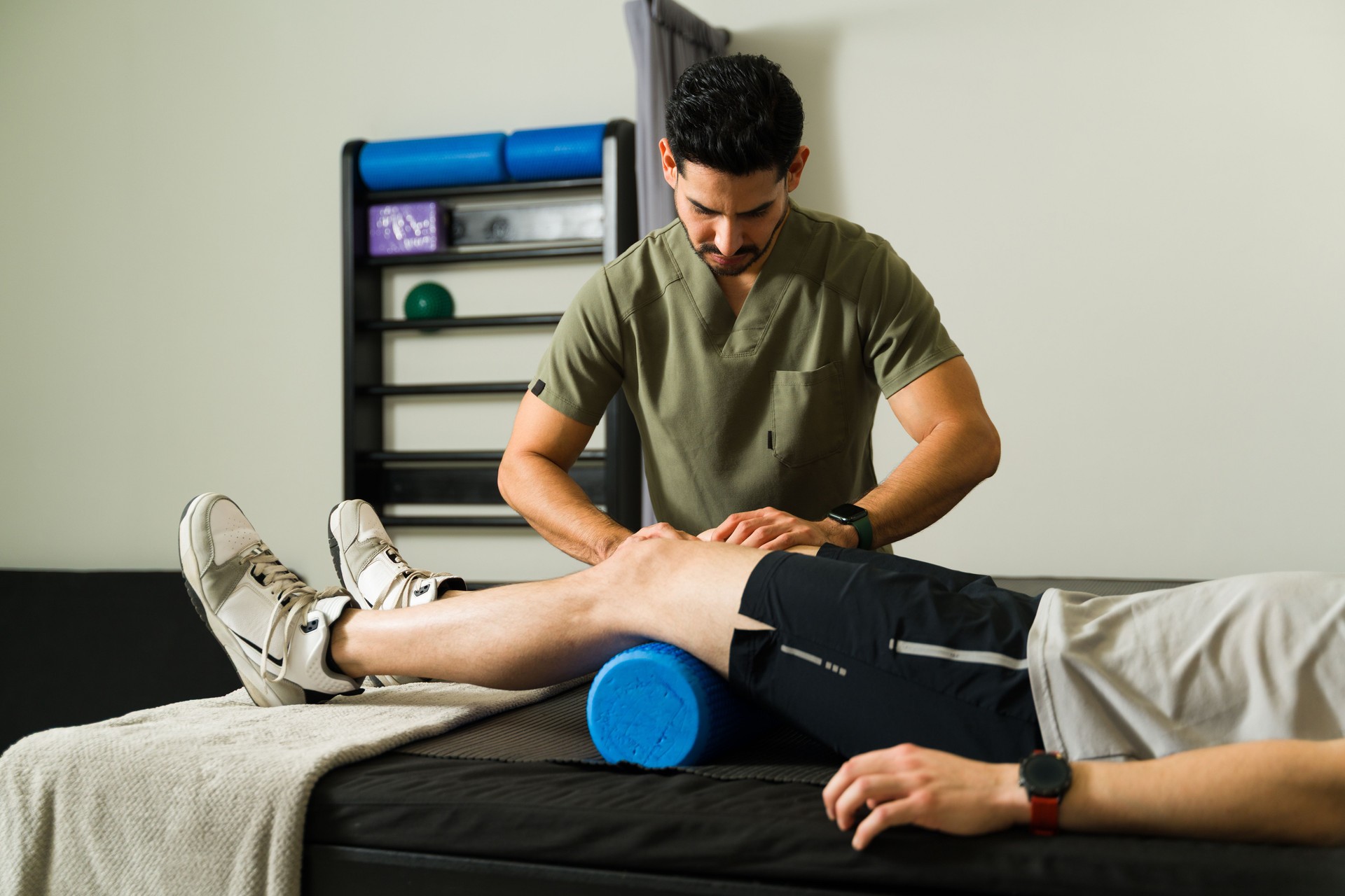 Professional physiotherapist performing knee massage using foam roller during sports rehabilitation treatment in modern medical facility